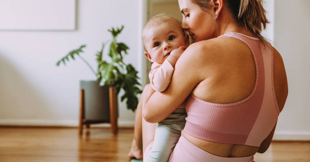 A mother wearing workout clothes holds her baby. The two are in their home and sitting on a workout mat.