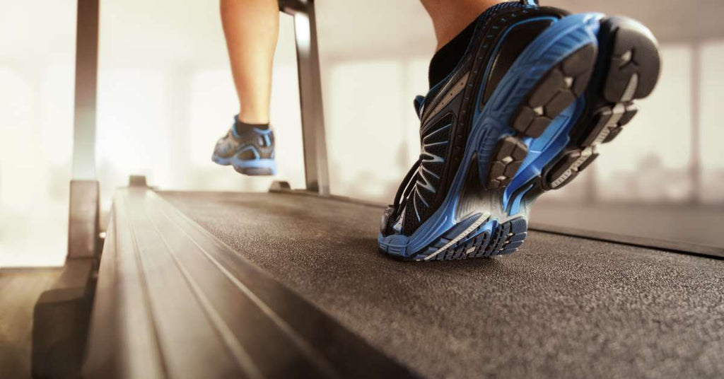 A person wearing green and black shoes runs on a treadmill. The treadmill has a black frame around the belt.