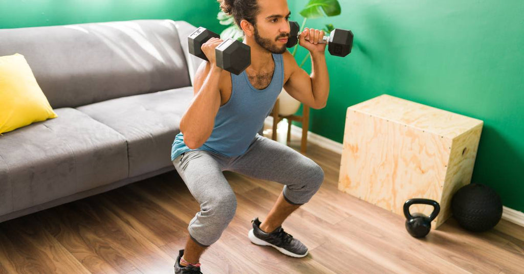 A man works out in his home, doing squats with dumbbells. His home has a gray couch and green walls.