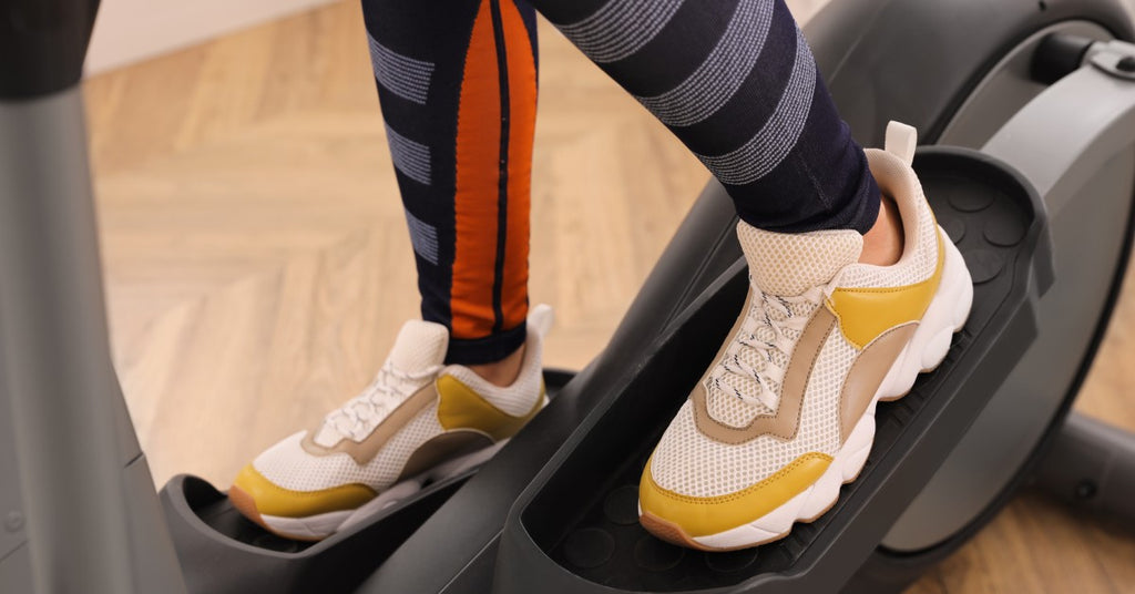 A woman's legs operate the pedals of her elliptical machine. She wears yellow, white, and tan sneakers.