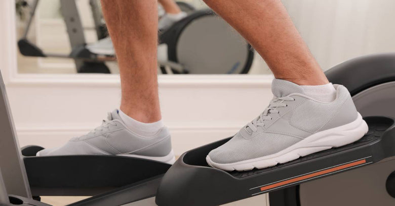 A man's feet move in the pedals of an elliptical machine as he exercises. A mirror stands behind him.