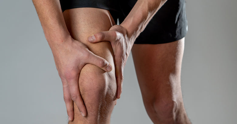 A man's hands hold his right knee as if it were giving him pain. He wears black shorts and is in front of a white background.