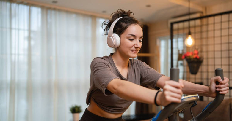 A woman smiles as she rides on her exercise bike in her home. She wears white over-the-ears headphones.