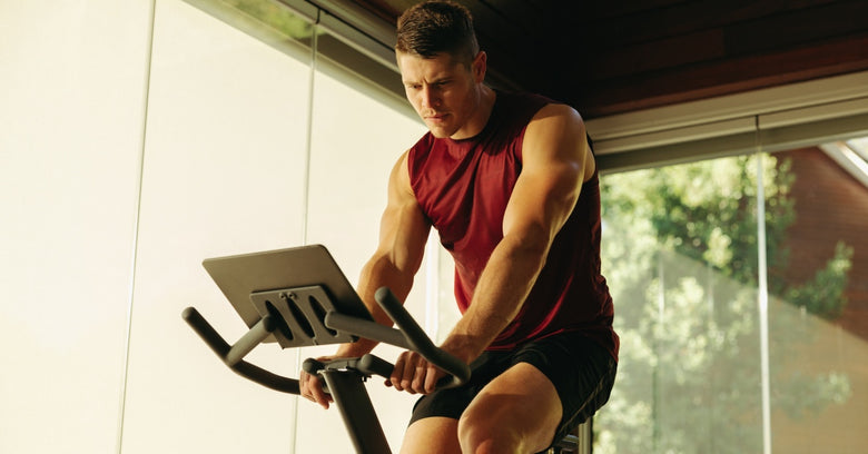 A man works out at home while riding his exercise bike. He wears a red shirt and black shorts and has short hair.